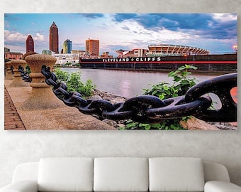 May include: A close-up of a black chain link fence with a view of the Cleveland skyline and a large ship with the text "CLEVELAND-CLIFFS" on the side. The skyline includes tall buildings and a stadium.