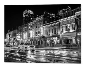 May include: A black and white photograph of a city street at night with a taxi cab driving down the street. The street is wet from rain and the buildings are lit up. The sign for the Hilton hotel is visible in the background.