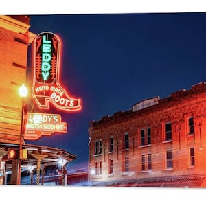 May include: A neon sign in the shape of a boot with the text "Leddy's Hand Made Boots" in red and green. The sign is mounted on the side of a brick building with a wooden awning. The building is lit up at night.