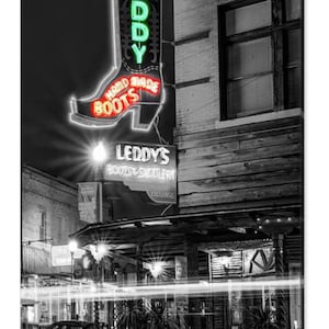 May include: A black and white photo of a storefront with a neon sign that reads "Leddy's Boots & Saddlery Exchange". The sign is shaped like a cowboy boot with the words "Hand Made Boots" in red.