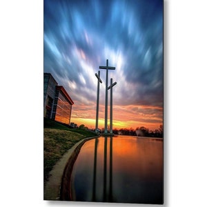 May include: Three large white crosses stand tall against a dramatic sunset sky with streaks of blue, pink, and orange clouds. The crosses are reflected in a still pond in front of a modern building.