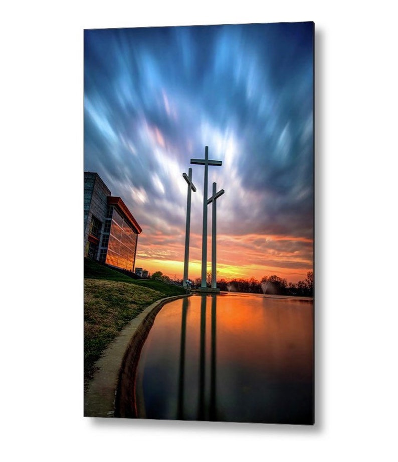 May include: Three large white crosses stand tall against a dramatic sunset sky with streaks of blue, pink, and orange clouds. The crosses are reflected in a still pond in front of a modern building.