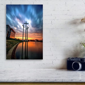 May include: Three large crosses stand tall against a dramatic sunset sky with clouds. The crosses are reflected in a still body of water. A building is visible in the background.