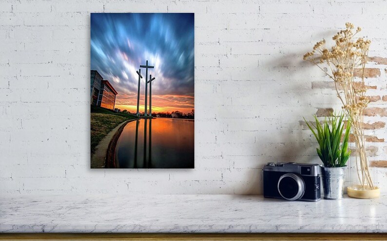 May include: Three large crosses stand tall against a dramatic sunset sky with clouds. The crosses are reflected in a still body of water. A building is visible in the background.