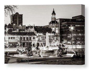May include: A black and white photograph of a fountain in a city square at night. The fountain is surrounded by buildings, including a tall building with a clock tower. The fountain is lit up, and the water is spraying high into the air.