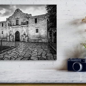 May include: A black and white wall art print depicting the Alamo mission building with its stone facade and arched entrance, set against a cloudy sky. The scene features a stone-paved courtyard in the foreground, capturing the historic architecture.