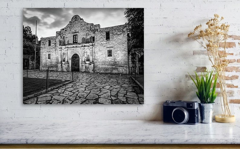 May include: A black and white wall art print depicting the Alamo mission building with its stone facade and arched entrance, set against a cloudy sky. The scene features a stone-paved courtyard in the foreground, capturing the historic architecture.