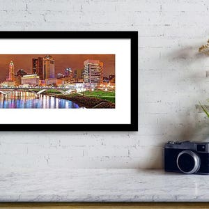 May include: A panoramic view of the Columbus, Ohio skyline at night. The cityscape is illuminated with lights reflecting in the river below. The buildings are tall and modern, with a mix of styles. The river is calm and still, reflecting the city lights.