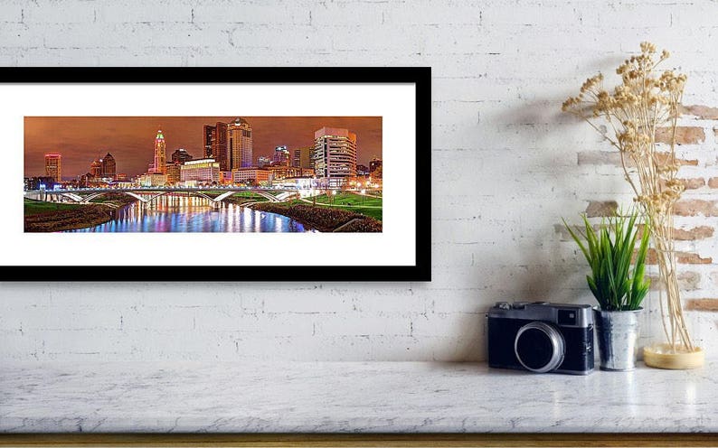 May include: A panoramic view of the Columbus, Ohio skyline at night. The cityscape is illuminated with lights reflecting in the river below. The buildings are tall and modern, with a mix of styles. The river is calm and still, reflecting the city lights.