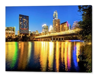 May include: A night view of the Austin skyline with a bridge in the foreground. The city lights are reflected in the water. The buildings are illuminated in various colors, including blue, yellow, and red.