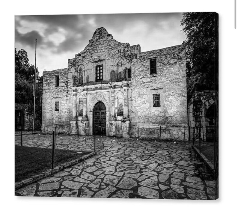May include: A black and white photograph of the Alamo mission building, featuring its iconic stone facade, arched wooden doorway, and rectangular windows. The structure is set behind a low metal fence with a stone-paved courtyard in the foreground.