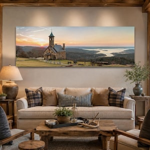 May include: A panoramic wall art print featuring a stone church with a steeple situated on a grassy hill overlooking a lake at sunset. The landscape scene displays warm orange and yellow sky tones, rolling hills, and a calm body of water.