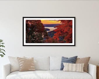 May include: A framed landscape photograph featuring a scenic view of a river and trees. The trees on the right side have vibrant red leaves, while the sky displays a gradient of orange and yellow hues. The photograph is displayed above a white sofa with pillows.