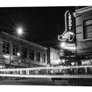 May include: Black and white photograph of a street scene at night with a neon sign for "Leddy's Boots" in the shape of a boot. The sign is lit up and the street is empty.