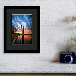 May include: A framed photograph of three crosses silhouetted against a dramatic sunset sky reflected in a still body of water.