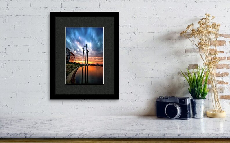 May include: A framed photograph of three crosses silhouetted against a dramatic sunset sky reflected in a still body of water.