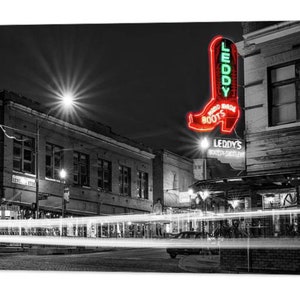 May include: A black and white photograph of a city street at night with a neon sign for "Leddy's Boots" in the shape of a red cowboy boot. The sign reads "Hand Made Boots".