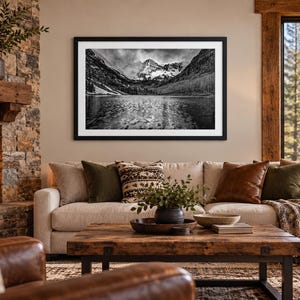 May include: A black and white framed landscape photograph depicting a mountain range reflected in a calm lake, mounted on a wall above a beige sofa with various throw pillows and a wooden coffee table in a living room setting.