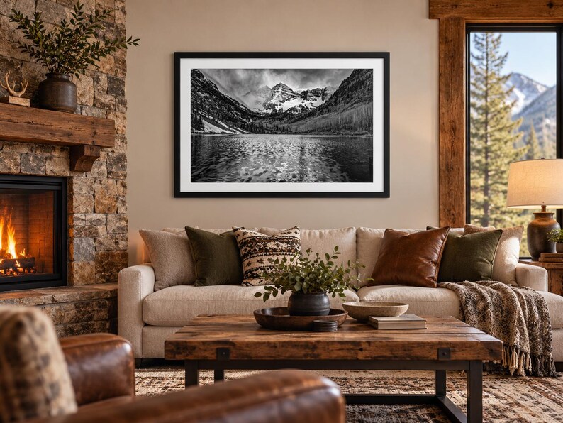 May include: A black and white framed landscape photograph depicting a mountain range reflected in a calm lake, mounted on a wall above a beige sofa with various throw pillows and a wooden coffee table in a living room setting.