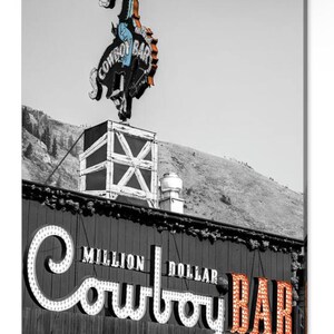 May include: A black and white photo of a building with a neon sign that says "Million Dollar Cowboy Bar". The sign is lit up in orange and white. A cowboy riding a bucking horse is on top of the building.