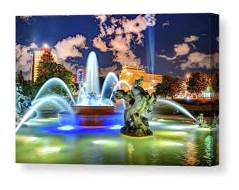 May include: A large fountain with multiple jets of water illuminated in blue and white light. The fountain is surrounded by trees and buildings, including a tall building with a clock tower. The fountain features a sculpture of a horse and rider.