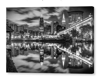 Pode incluir: Uma fotografia a preto e branco do horizonte de Cleveland à noite, com os arranha-céus da cidade refletidos na água. Os edifícios são iluminados por candeeiros de rua e pelo reflexo da lua.