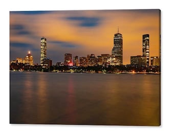 May include: A panoramic view of a city skyline at dusk. The city is lit up with lights, and the sky is a soft orange and pink. The water in the foreground is calm and reflective.