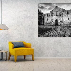 May include: A large black and white wall art print of the Alamo mission building, featuring a stone facade and a cobblestone courtyard, displayed in a room with a bright yellow armchair, a small wooden side table, and a modern pendant light.
