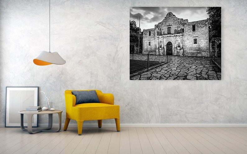 May include: A large black and white wall art print of the Alamo mission building, featuring a stone facade and a cobblestone courtyard, displayed in a room with a bright yellow armchair, a small wooden side table, and a modern pendant light.