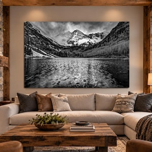 May include: A large rectangular black and white wall art print depicting a mountain range reflected in a calm lake. The landscape features snow-capped peaks, dense forest slopes, and a textured water surface, mounted above a beige sectional sofa.