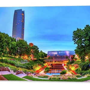 May include: A view of a modern glass building with a reflecting pool in front of it. The building is surrounded by lush green trees and landscaping. The sky is a light blue with some clouds.
