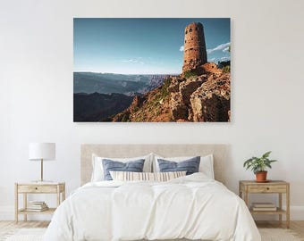 May include: A scenic landscape photograph of the Grand Canyon featuring a stone tower on a rocky outcrop. The tower is brown and cylindrical, set against a blue sky. The canyon's layered rock formations are visible in the background.