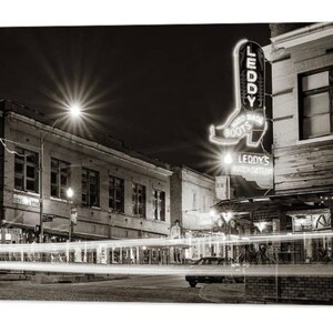May include: Black and white photograph of a city street at night. A neon sign in the shape of a cowboy boot reads "LEDDY'S BOOTS". Buildings line the street, and streaks of light from passing vehicles are visible.