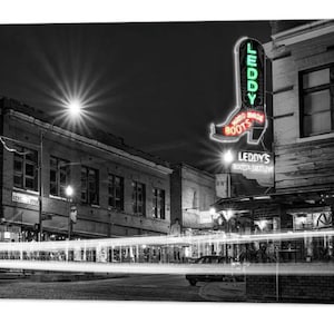 May include: Black and white photograph of a street scene at night with a neon sign for "Leddy's Boots" in the shape of a cowboy boot. The sign is lit up in red and green neon. The street is empty except for a few cars driving by. The buildings are brick and have windows. The street is lit by streetlights.