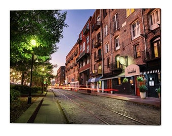 May include: A brick building with a cobblestone street in front of it. The street has streetlights and a row of trees on the left side. The building has a sign that says "1748", and there are lights coming from the windows.