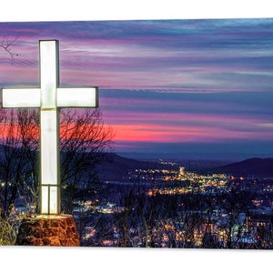 May include: A large white cross illuminated against a colorful sunset sky. The cross is silhouetted against a cityscape with twinkling lights in the distance.