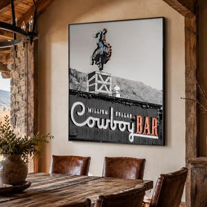 May include: A square wall art print featuring a black and white photograph of the Million Dollar Cowboy Bar sign. The image displays a neon cowboy figure atop a building, with the text Million Dollar Cowboy Bar in large, illuminated lettering.