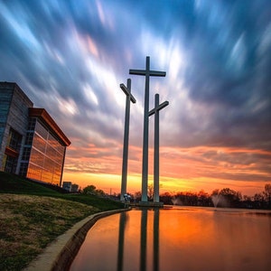 May include: Three large white crosses stand tall against a dramatic sunset sky with streaks of blue and orange clouds. The crosses are reflected in a still pond of water in front of a modern building.