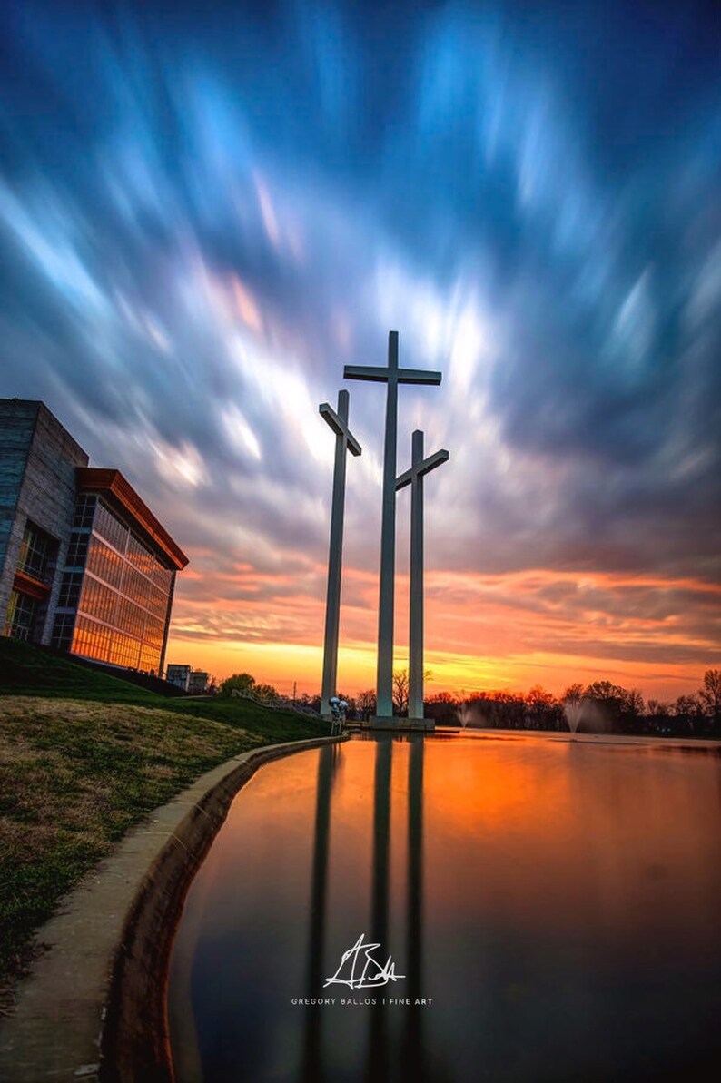 May include: Three large white crosses stand tall against a dramatic sunset sky with streaks of blue and orange clouds. The crosses are reflected in a still pond of water in front of a modern building.