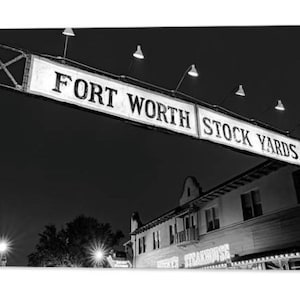 May include: Black and white photo of a sign that reads "Fort Worth Stock Yards" at night. The sign is illuminated by streetlights.
