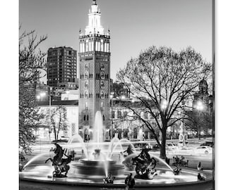 May include: A black and white photograph of a fountain in front of a building with a tall tower. The fountain has multiple water jets and sculptures of horses. The building has a tower with a pointed roof and a clock face.
