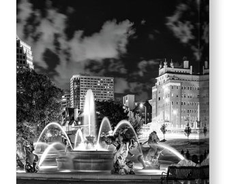 Puede incluir: Una fotografía en blanco y negro de una fuente en un parque de la ciudad por la noche. La fuente está rodeada de árboles y edificios. El agua está iluminada por luces, creando un hermoso efecto.