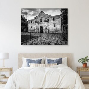 May include: A black and white wall art print depicting the historic Alamo mission building in San Antonio, Texas. The image features the stone facade of the structure, a cobblestone courtyard in the foreground, and a cloudy sky above the building.