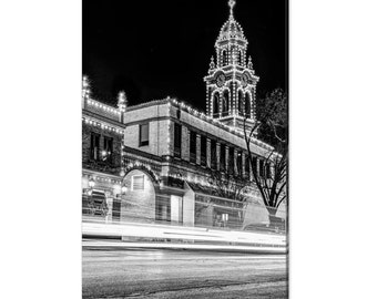 May include: A black and white photograph of a building with a clock tower at night. The building is decorated with string lights and there are cars driving by in the street.