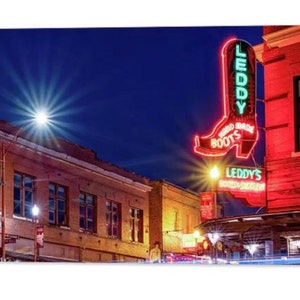 May include: A neon sign for "Leddy's Boot Shop" in the shape of a cowboy boot, with the words "World Made Boots" on the boot. The sign is lit up in red and green neon, and is mounted on the side of a brick building. The building is lit up with yellow lights, and there are other buildings in the background.