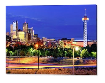 May include: A nighttime cityscape with a tall tower in the foreground and a skyline of skyscrapers in the background. The tower is lit up with white lights and has a red roof. The skyscrapers are also lit up with lights, and there are trees and a highway in the foreground.