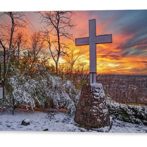 May include: A white cross stands tall on a stone base, covered in snow, against a backdrop of a vibrant orange and pink sunset. The cross is silhouetted against the sky, with trees in the foreground and a cityscape in the distance.
