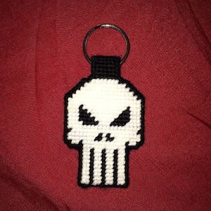 May include: A white and black skull keychain with a silver key ring. The skull is made of plastic canvas and has a pixelated design.