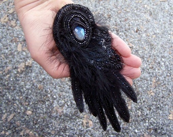 May include: A black feather brooch with a large iridescent blue gemstone in the center. The brooch is surrounded by black beads and sequins.