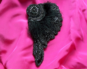 May include: A black beaded and feathered brooch in the shape of a wing. The brooch is on a pink fabric background.